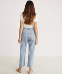 A woman with long brown hair stands facing away, wearing a white t-shirt, 90s Crop Jean in light blue denim, and sandals against a plain light background.