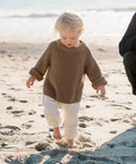 A young child with blonde hair, barefoot in the sand by the ocean, wears a Kids Cotton Fisherman sweater and white pants.