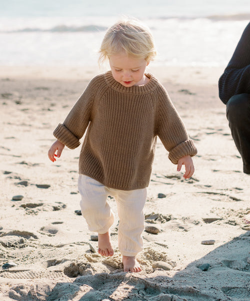 A young child with blonde hair, barefoot in the sand by the ocean, wears a Kids Cotton Fisherman sweater and white pants.