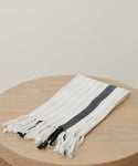 The Striped Hand Towel, featuring thin beige stripes, a bold black stripe, and fringed edges, is displayed folded on a round wooden surface against a simple light background.