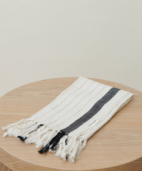 The Striped Hand Towel, featuring thin beige stripes, a bold black stripe, and fringed edges, is displayed folded on a round wooden surface against a simple light background.