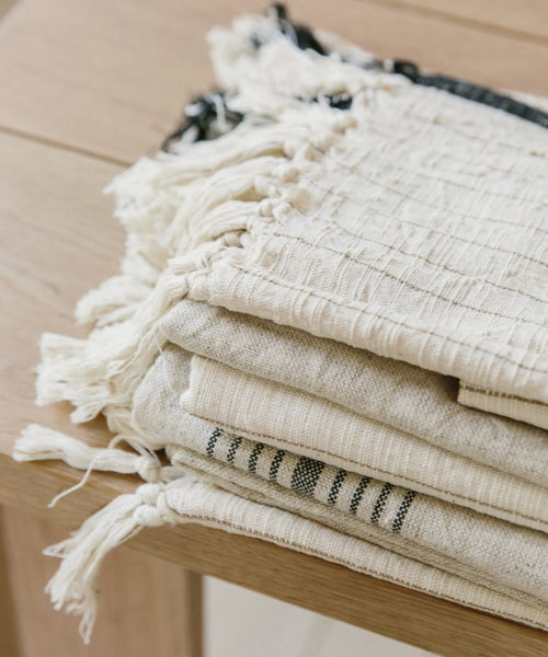 A close-up of several neatly folded, off-white and beige textured blankets with fringed edges stacked on a light wooden surface. Among them is the Striped Hand Towel, featuring a classic black stripe and subtle patterns.