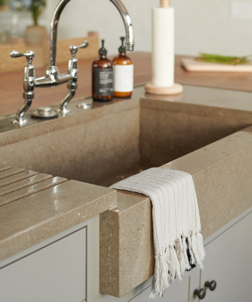 A modern farmhouse kitchen sink features a beige stone basin, silver faucet, and a Striped Hand Towel with black stripes draped over the front, along with two brown soap dispensers on the counter in the background.