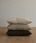 The Luna Lumbar Pillow, handwoven in neutral shades of beige, light brown, and dark brown, is stacked with two other textured throw pillows on a light wooden floor against an off-white wall.