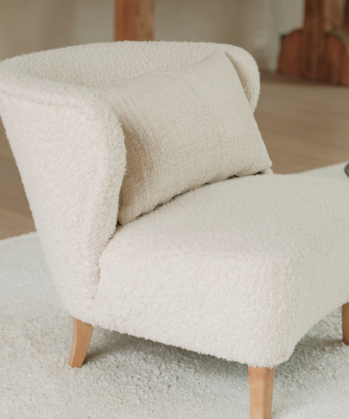 A modern, cream accent chair with wooden legs sits on a light rug in a cozy, minimalist room, complemented by the Luna Lumbar Pillow—handwoven and filled with a feather down insert.