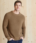A man with short brown hair, smiling with his hands in his pockets, wears the Mens Cotton Fisherman sweater featuring ribbed edge detailing and dark jeans while standing against a light wooden wall.