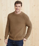 A man wearing the Mens Cotton Fisherman sweater and blue jeans stands in front of a light wooden wall, smiling with one hand in his pocket.