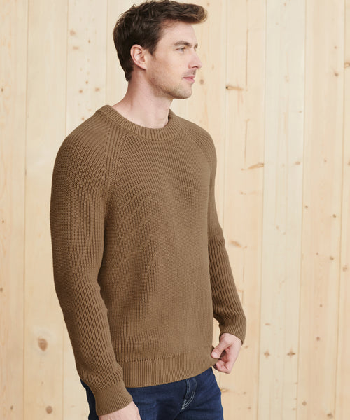 A man in a Mens Cotton Fisherman sweater and dark blue jeans stands sideways before a light wooden wall, showing ribbed edge details and a neutral expression.