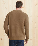 A man, seen from behind, stands facing a light wood wall wearing the Mens Cotton Fisherman sweater with ribbed edges and dark blue jeans. His left hand is slightly bent.