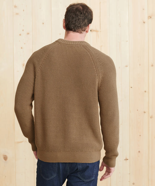 A man, seen from behind, stands facing a light wood wall wearing the Mens Cotton Fisherman sweater with ribbed edges and dark blue jeans. His left hand is slightly bent.
