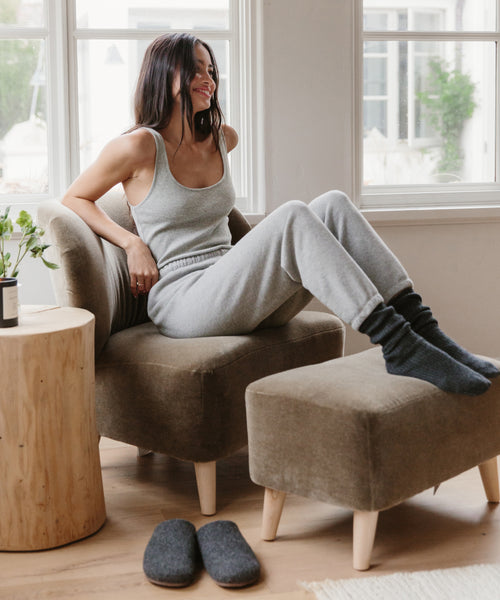 A woman in Saturday Sweatpant and a grey tank top lounges with her feet up on an ottoman, smiling, as slippers rest nearby in a bright, sunlit room.