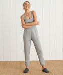 A smiling woman stands on a woven rug in front of a white paneled wall, wearing a grey tank top and the ultra-soft Saturday Sweatpant. Her arms are crossed, her hair is tied back, and she completes the look with dark grey slippers.