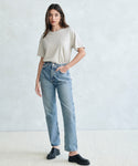 A woman stands against a light gray textured wall, wearing the Vintage Pocket Tee tucked into high-waisted blue jeans and black shoes—a relaxed, casual style ideal for everyday wear.