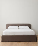 The Cove Bed features a minimalistic design with a gray upholstered headboard and base, dressed in white bedding and two white pillows, positioned against a plain white wall on a light wooden floor.