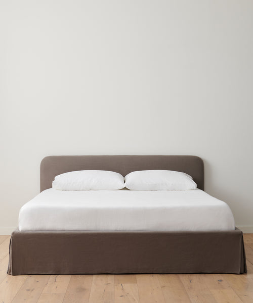The Cove Bed features a minimalistic design with a gray upholstered headboard and base, dressed in white bedding and two white pillows, positioned against a plain white wall on a light wooden floor.