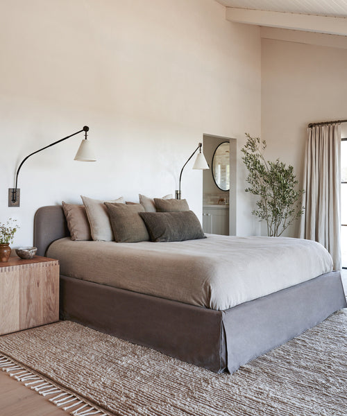 A minimalist bedroom with a large bed dressed in the Cove Bed Slipcover in Charcoal Linen, neutral bedding, wall-mounted lamps, a wooden nightstand, textured rug, potted plant, and soft natural light through beige curtains.