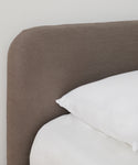 A close-up of a bed featuring the Cove Bed Slipcover, paired with a brown upholstered headboard and two stacked white pillows on Belgian Libeco Linen bedding against a plain white wall.