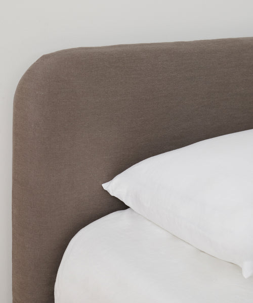 A close-up of a Cove Bed features a brown upholstered headboard and crisp white bedding, including a white pillow and sheets, set against a plain white wall.