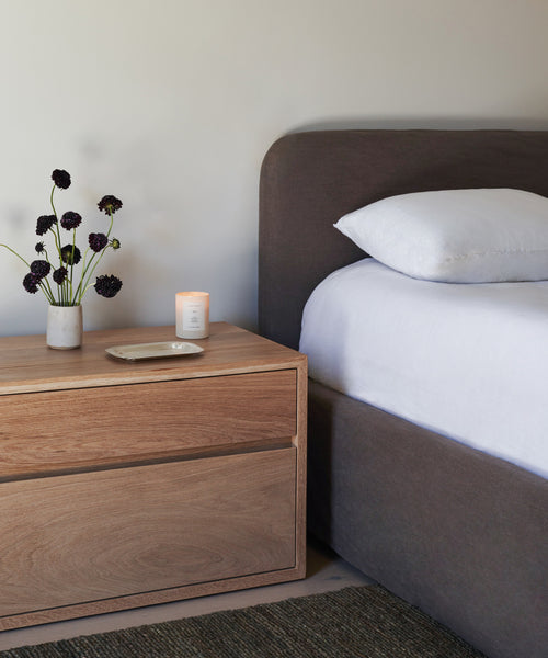 A wooden nightstand with a small vase of dark purple flowers, a lit candle, and a dish sits beside the bed with white bedding and an upholstered headboard, making it perfect to pair with the Cove Bed.