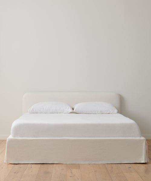 The Cove Bed, featuring an upholstered headboard, two white pillows, and a Belgian Libeco linen bedspread, stands against a plain off-white wall on a light wooden floor.