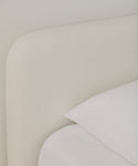 Close-up of the Cove Bed in ivory linen with a rounded, upholstered headboard and white Belgian Libeco linen bedding, highlighting its soft, minimalist design against a light wall.