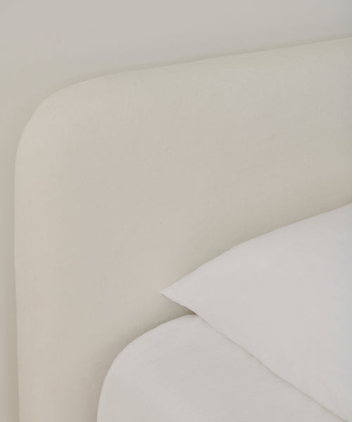 Close-up of a bed featuring the Cove Bed Slipcover in soft, light beige upholstery with crisp white Belgian Libeco linen bedding, neatly arranged pillow and sheet, for a minimalistic and modern look.