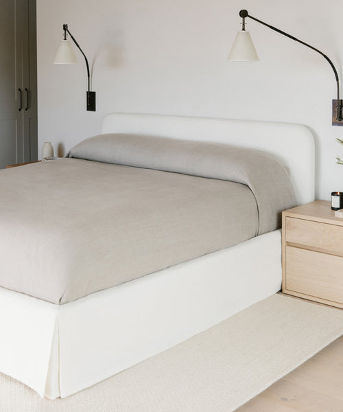 A minimalist bedroom with a neatly made bed featuring the Cove Bed Slipcover and a white headboard. Two black wall-mounted lamps flank the bed, while a light wood nightstand sits on the right side.