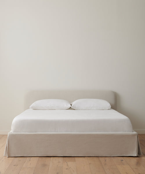 The Cove Bed Slipcover features a light beige upholstered headboard and base, styled with natural linen bedding and two white pillows, positioned against an off-white wall on a wooden floor for a minimalist look.