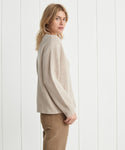 A blonde woman stands sideways against a white paneled wall, looking over her shoulder at the camera while wearing the Cropped Cashmere Cocoon Cardigan in oatmeal and brown pants.
