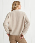 A person with blonde hair, seen from behind, is wearing the Cropped Cashmere Cocoon Cardigan in cream with tan pants, standing before a white paneled wall.