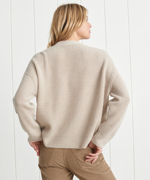 A person with blonde hair, seen from behind, is wearing the Cropped Cashmere Cocoon Cardigan in cream with tan pants, standing before a white paneled wall.