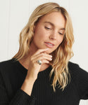 A woman with wavy blonde hair wears a black sweater and the Felix Dome Ring in white gold, gently resting her hand near her chin. The background is light and minimal.