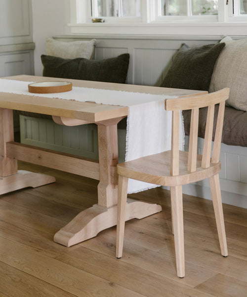 A light wooden table with a matching chair sits on a wooden floor. The table is topped with the Frayed Linen Runner and a round tray. Behind it, a built-in bench with gray and beige cushions rests under the window.