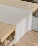 A close-up of a light wooden table showcases the Frayed Linen Runner, neatly draped over the edge. Its white, textured fabric and fringed edges highlight the runners simple, elegant design and soft linen material.