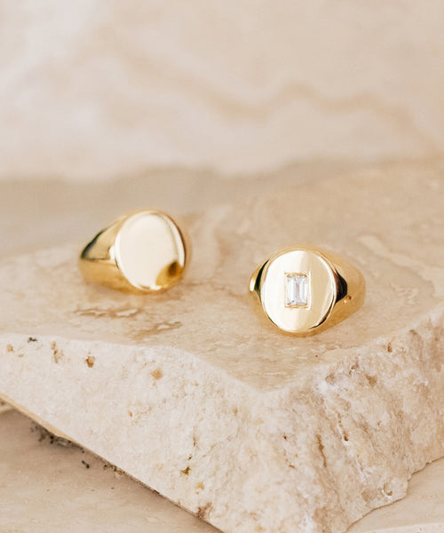 Two gold signet rings rest on a beige stone surface: one is the Gemma Baguette Signet Ring from Jenni Kayne Jewelry with a rectangular gemstone, and the other is a smooth, plain 14k gold ring.