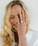 A smiling woman with long blonde hair covers one eye with her hand, highlighting several gold rings, including the Gemma Baguette Signet Ring. She wears a textured top against a soft, pale background.