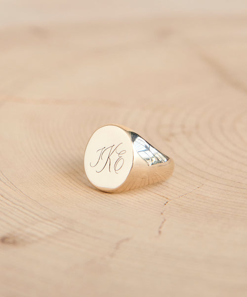 A gold signet ring with engraved initials JKC in elegant script rests on a light wooden surface—a timeless piece, expertly customized with our Custom Engraving Service.