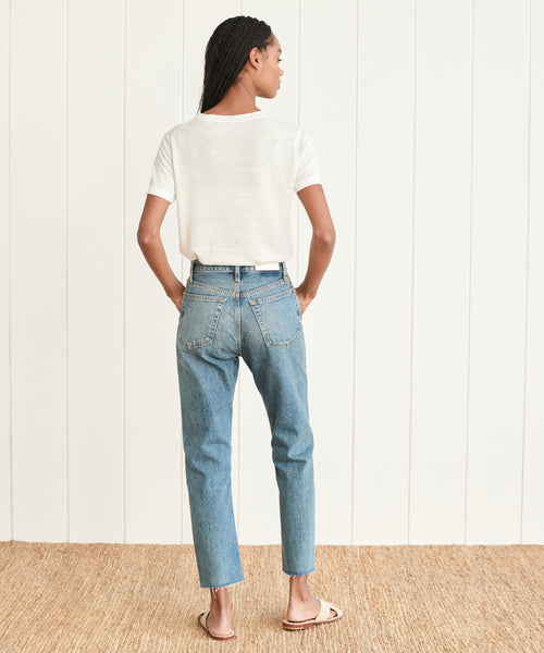 A woman stands facing away, wearing a white short-sleeve t-shirt, High Rise Stove Pipe Jean, and sandals on a woven rug in front of a white paneled wall.