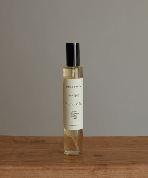 A clear glass bottle of Mandeville Room Spray with a black cap rests on a wooden surface against a gray background. The label features clean, nature-inspired scents of lemon, eucalyptus, and vetiver from Los Angeles.
