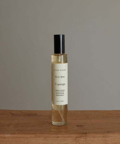 A clear glass bottle of Topanga Room Spray with a black cap sits on a wooden surface against a neutral background, evoking nature-inspired scents.