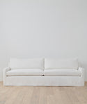 A simple, modern Harbor Sofa in ivory linen with two seat and two back cushions sits against a plain white wall on light wood flooring.