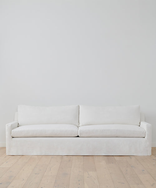 A simple, modern Harbor Sofa in ivory linen with two seat and two back cushions sits against a plain white wall on light wood flooring.
