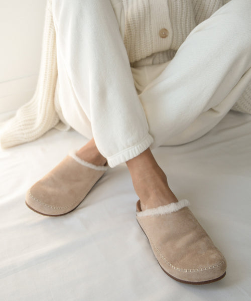 Dressed in cozy, cream loungewear, a person sits cross-legged on a white bed, wearing the Shearling-Lined Moc Clog—handmade in Italy with a cork sole and soft white fleece lining.