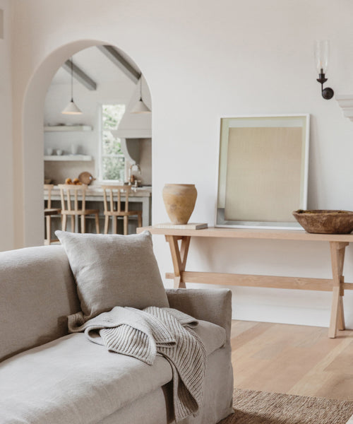A neutral-toned living room has a beige sofa with a knitted blanket, a wooden console table with decor, and the Untitled 2 Framed Print from Jenni Kayne Home, plus an arched doorway to a kitchen with light wood stools and pendant lights.