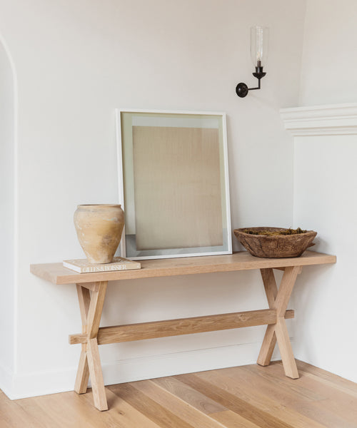 A wooden bench holds a tan vase on a book, the Untitled 2 Framed Print, and a rustic bowl with moss—styled with Jenni Kayne Home charm—against a white wall with a black sconce above and light wood flooring below.
