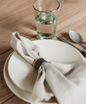 A set dining table features a white plate, beige cloth napkin held by the Ceramic Napkin Rings Set of 4, a glass of water, and a spoon and knife on a light wooden surface.
