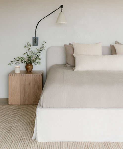 Minimalist bedroom featuring the Cove Bed in ivory linen with a beige upholstered headboard, neutral pillows, a wooden nightstand topped with a ceramic vase of greenery, candle, and modern wall lamp. Light textured rug on the floor.