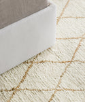 Close-up of the Wilder Diamond Jute Rug, a light-colored, handwoven rug featuring a tan diamond pattern beside a bed with a white upholstered frame and light bedding.