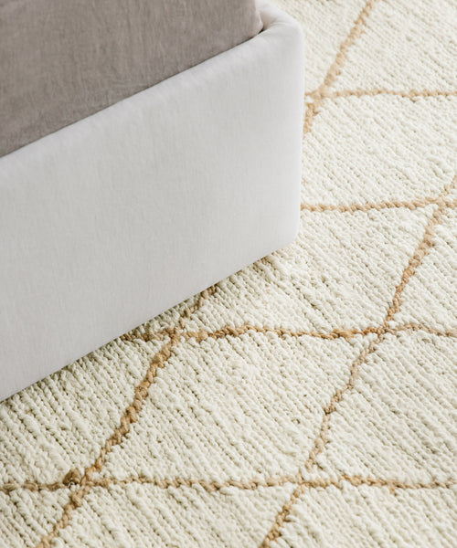 Close-up of the Wilder Diamond Jute Rug, a light-colored, handwoven rug featuring a tan diamond pattern beside a bed with a white upholstered frame and light bedding.