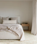Minimalist bedroom with a neatly made bed in neutral tones, beige bedding, and pillows. A wooden nightstand with a bowl and candle sits beside the bed. Soft light filters through curtains onto a Wilder Diamond Jute Rug.